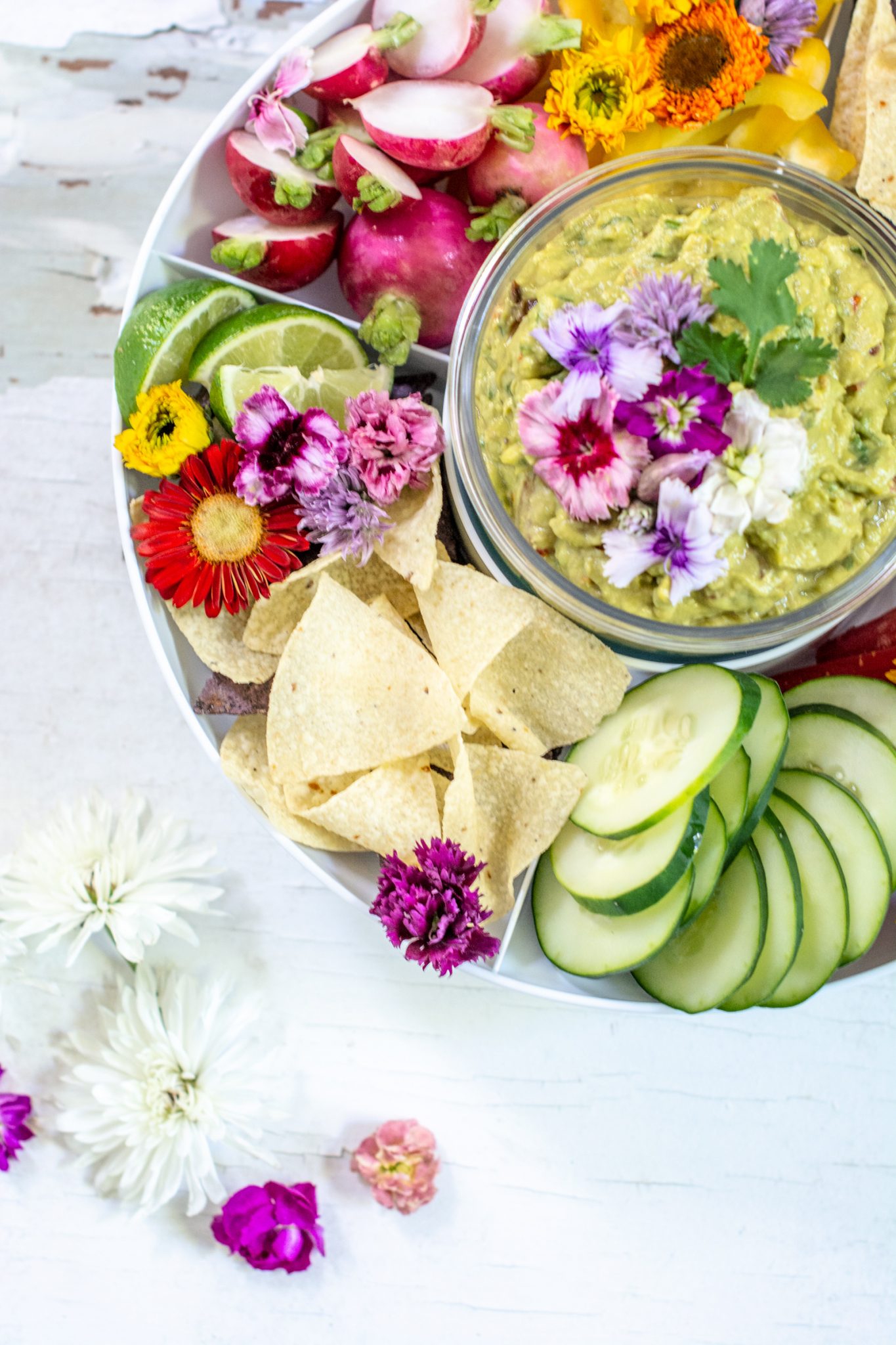 Fresh sliced vegetables surrounding a bowl of spicy guacamole on a white table.