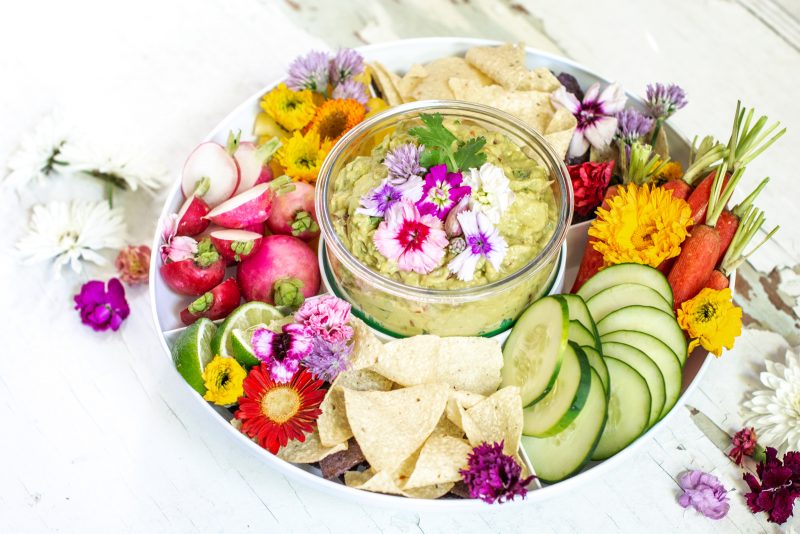 Overhead view of guacamole surrounded by fresh sliced vegetables and decorated with white and pink flowers on white background.