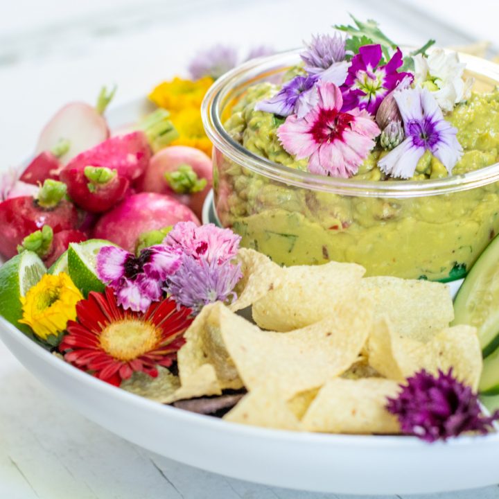 Spicy Guacamole recipe in a bowl served with fresh vegetables and tortilla chips on a white table.