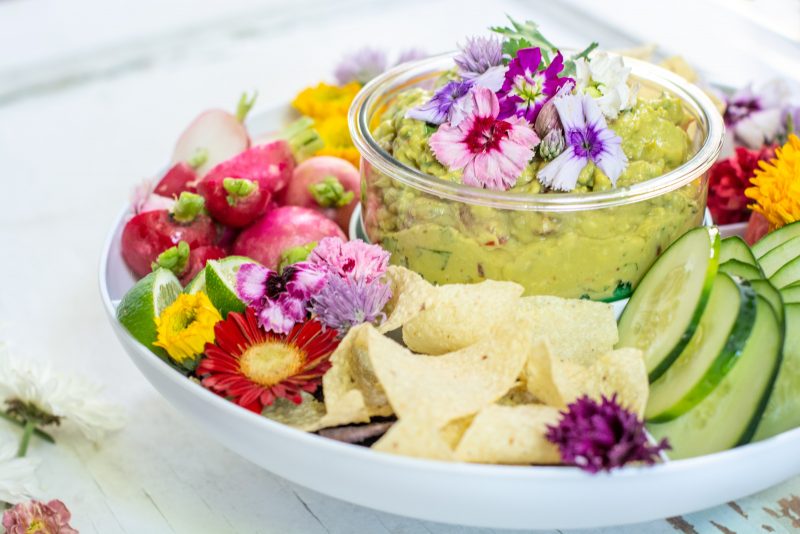 Spicy Guacamole recipe in a bowl served with fresh vegetables and tortilla chips on a white table.