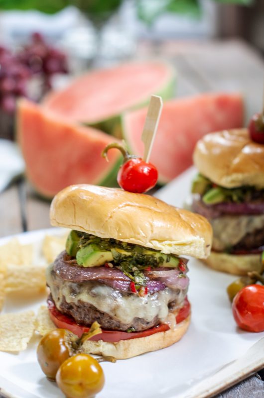 A chimichurri burger recipe with melted cheese, tomatoes, caramelized onions, avocado slices and cherry peppers