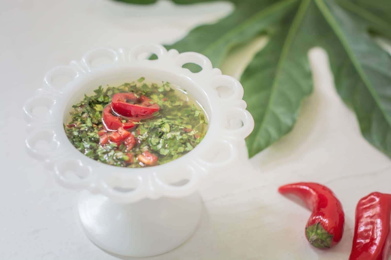 Chimichurri Sauce recipe in a white pedestal bowl with red chili peppers and greenery on white table