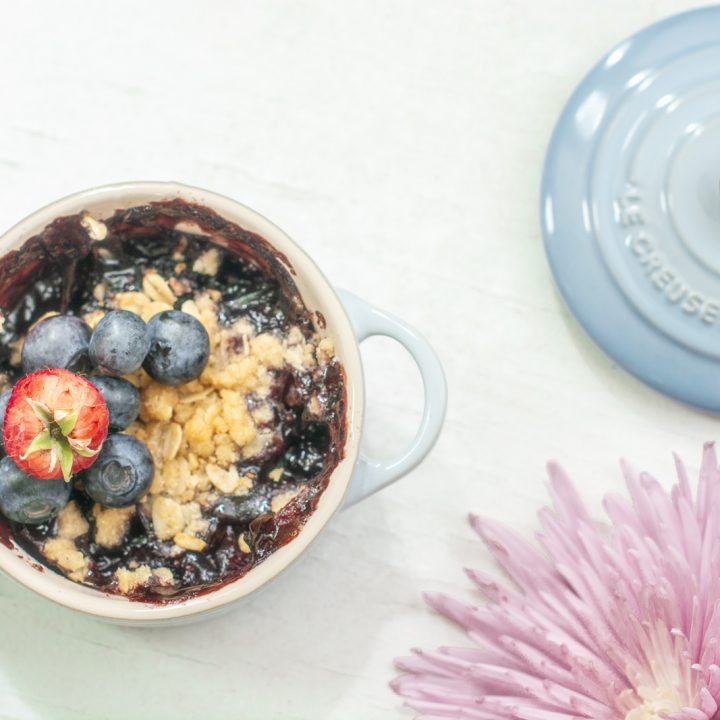A mixed berry cobbler dessert on a white background with pink flower and blue lid