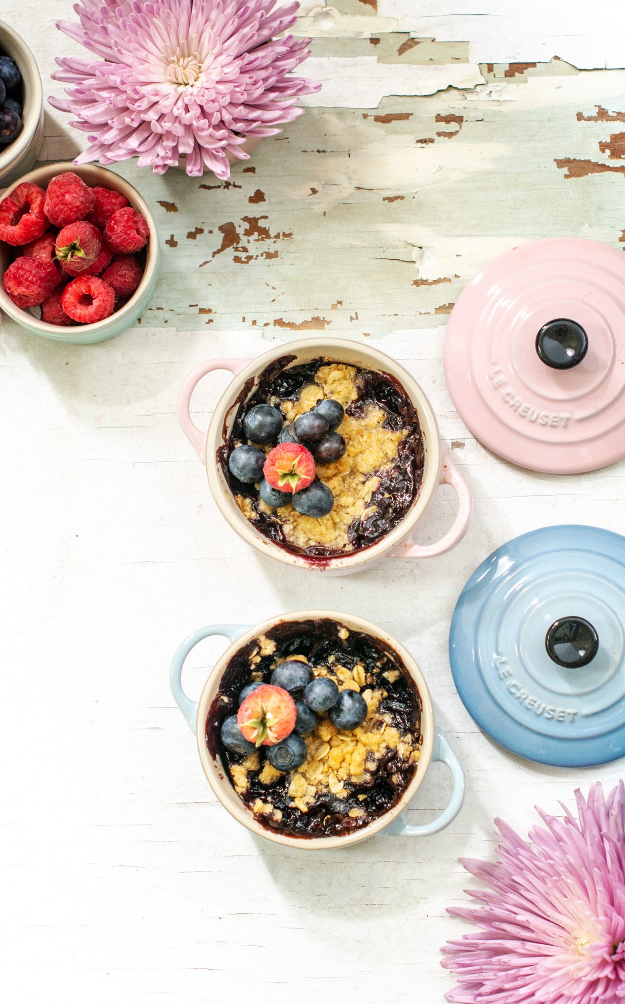 A raspberry and blueberry crisp recipe on a white table with pink and blue Le Creuset lids.