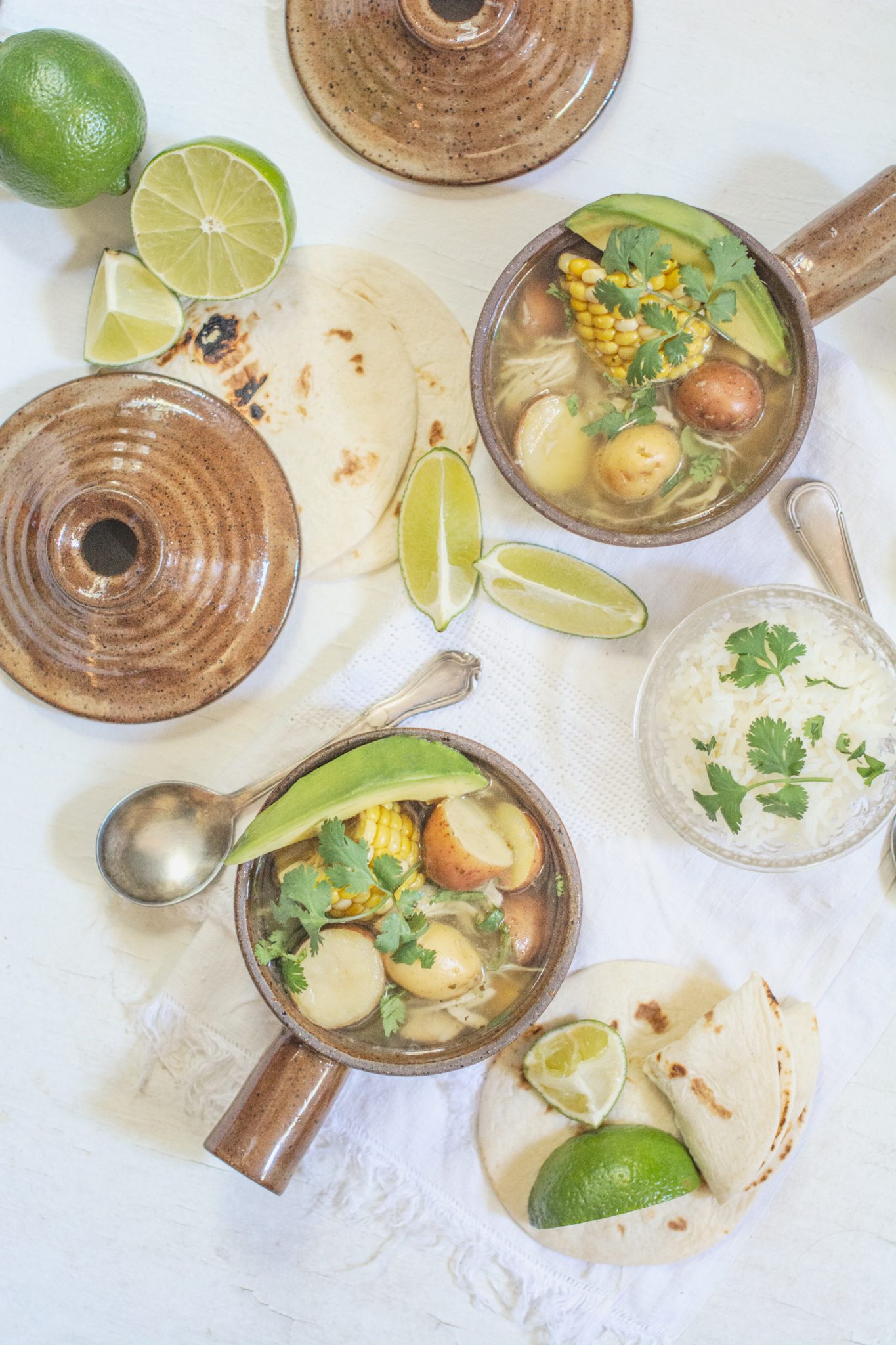 Ajiaco recipe; Colombian Chicken Corn and Potato Stew served on a white table with tortillas, limes and bowls of rice.